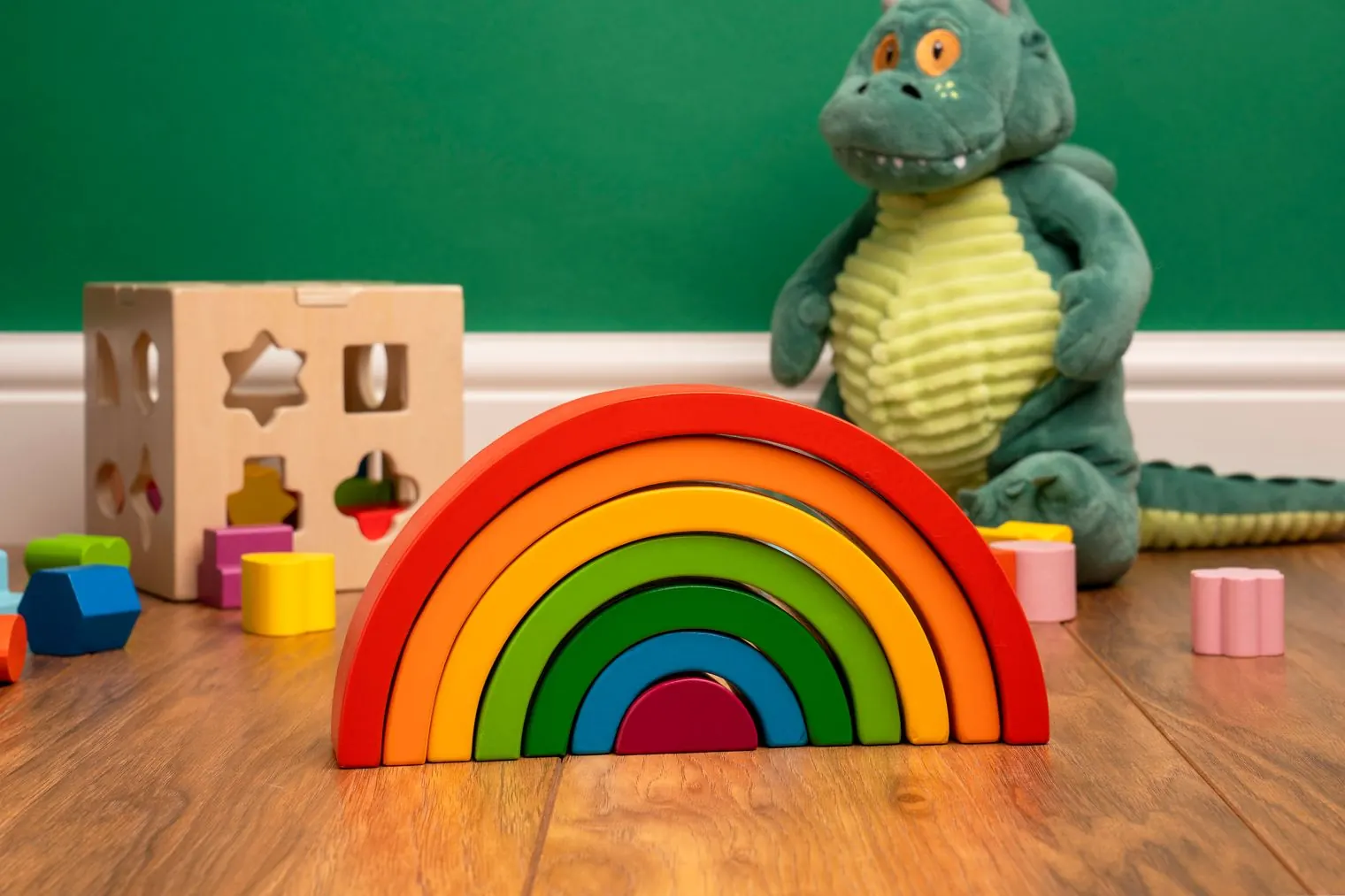 A wooden rainbow stacker, a shape sorter and a plush dinosaur on a wooden floor