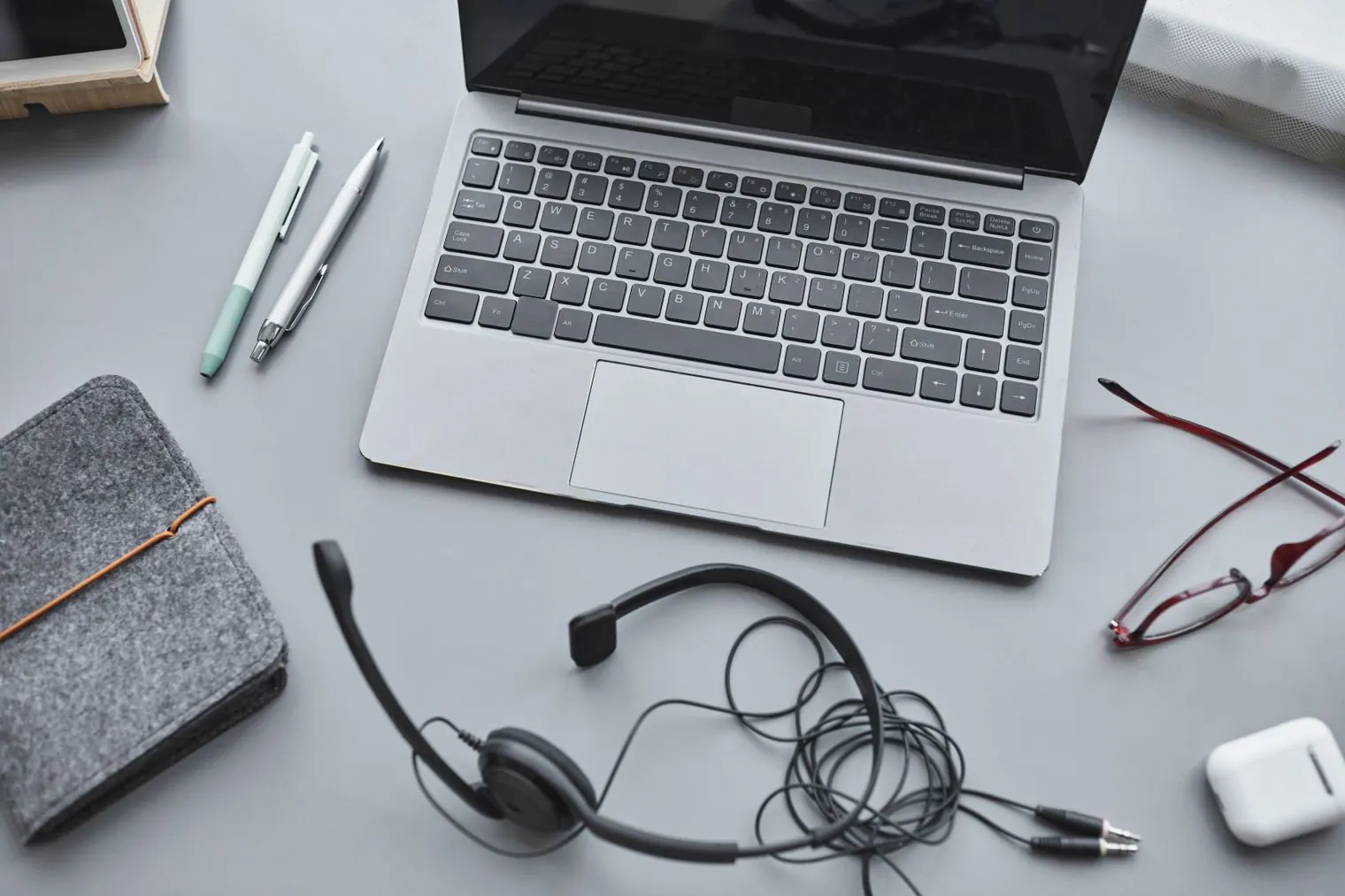 A laptop on a desk with a headset, pens, glasses and a notebook